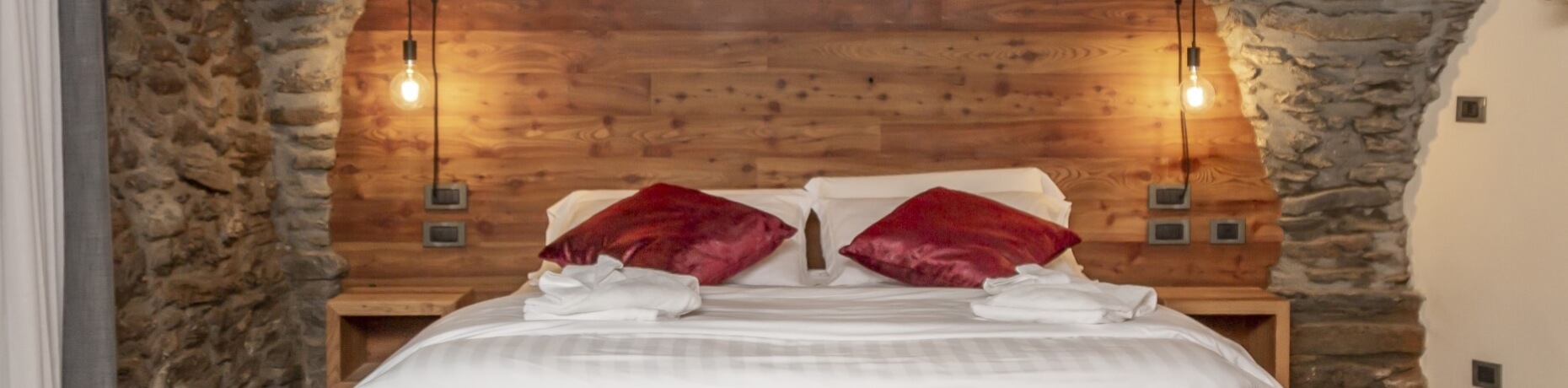 Serene bedroom with two beds, rustic stone wall, red pillows, stylish light fixtures, and wooden tables with charging ports.