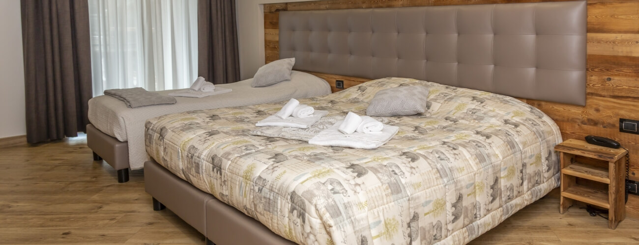 Hotel room with two beds, one with elephant print quilt, wooden nightstand, window, and light wood floor.