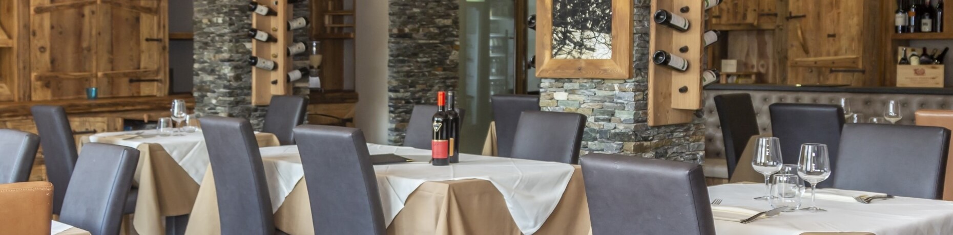 Upscale restaurant dining area with white tablecloths, wine glasses, and a stone wall featuring a wine rack and mirror.