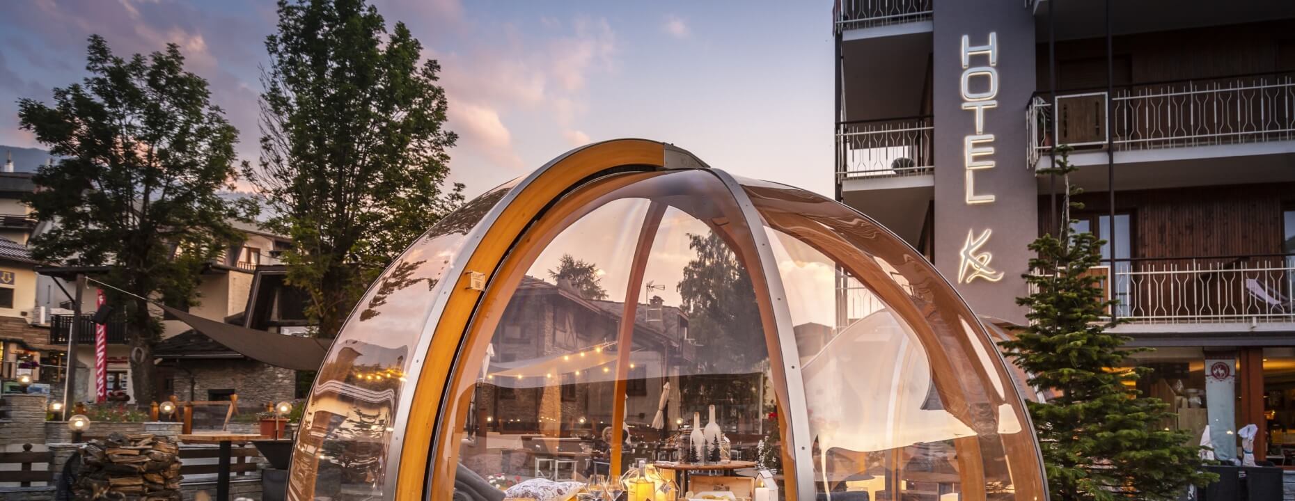 A transparent dome restaurant in front of a hotel, with wine bottles inside and a blue sky with pink clouds above.