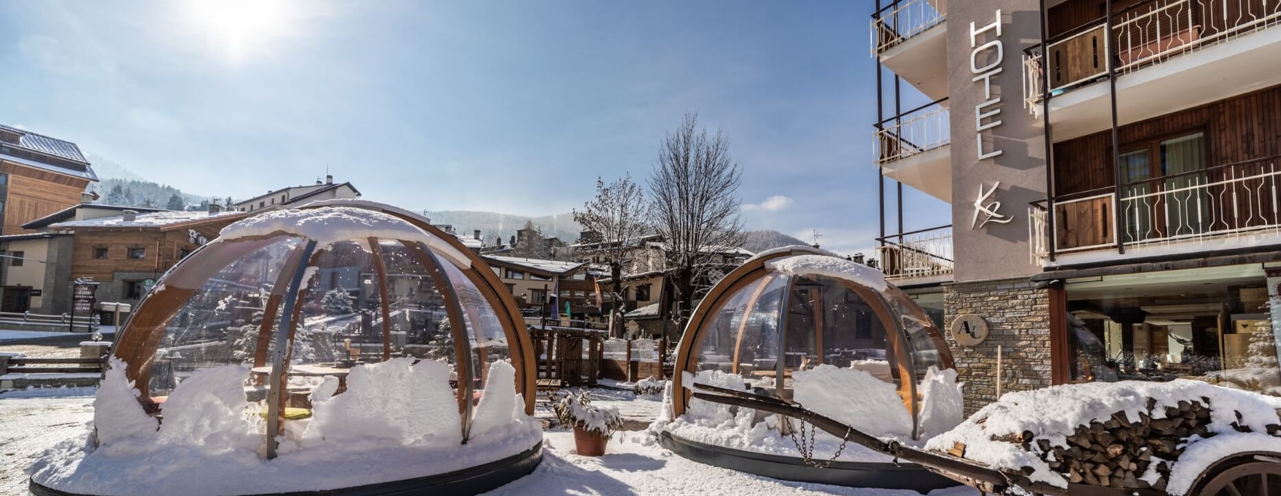 Due cupole trasparenti con tavoli, un hotel con facciata in pietra e legno, e un carrello di legna su un terreno innevato.