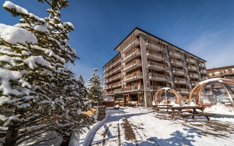 Serene winter scene featuring a multi-story building with balconies, snow-covered pine tree, and clear outdoor seating domes.