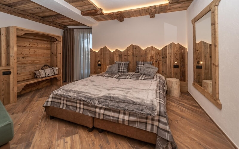 Cozy rustic bedroom with a central bed, wooden walls, a bench, a large mirror, and a window with curtains.