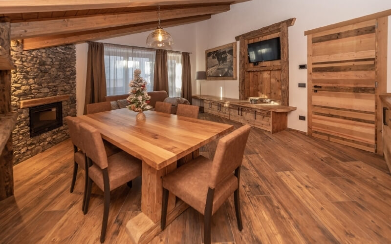 Cozy dining and living area with a wooden table, chairs, fireplace, couch, and Christmas tree, featuring white walls and hardwood floor.