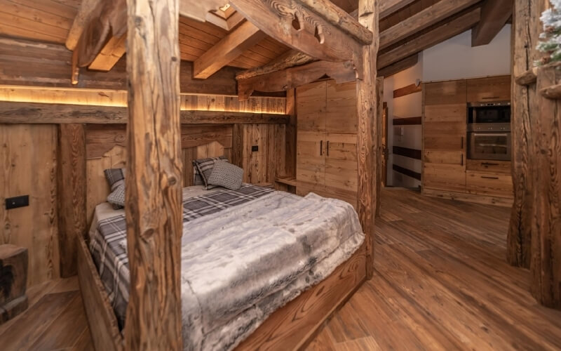 Camera da letto rustica con letto centrale, coperta grigia e cucina sullo sfondo con armadietti e forno.