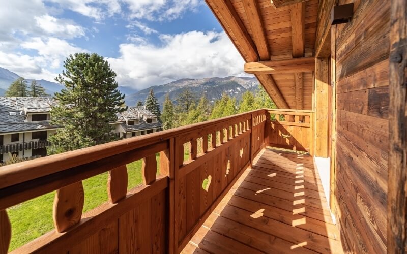 Wooden balcony with decorative railing overlooking snow-capped mountains and trees, bathed in warm sunlight.