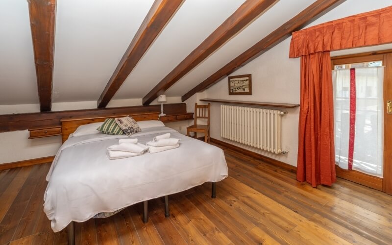 Cozy attic bedroom with a bed, chair, radiator, window with curtains, and wooden beams on light wooden floors.