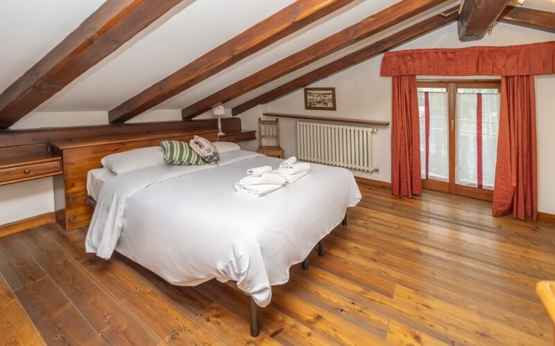 Cozy room with a bed, wooden floors, a window with red curtains, and a small table, well-lit by natural light.
