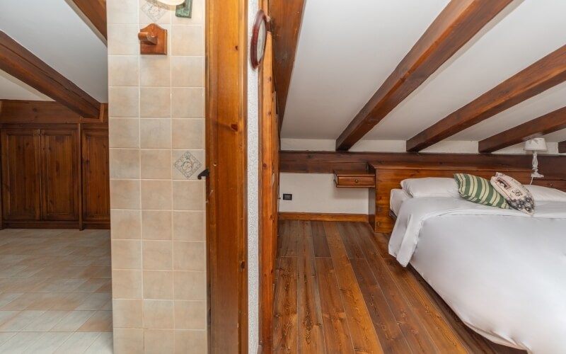 Cozy rustic bedroom with lofted ceiling, large bed, green striped pillow, and a shelf above, leading to a tiled bathroom.