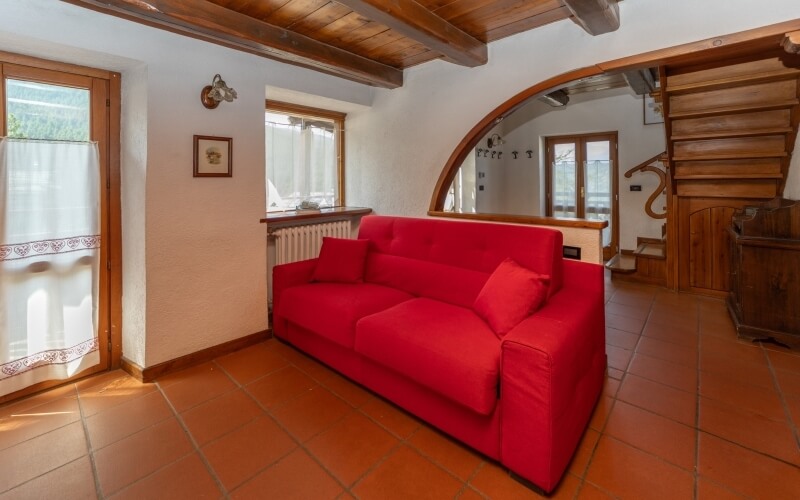Cozy living room featuring a plush red couch, wooden staircase, doorway, large window, and light brown tile floor.