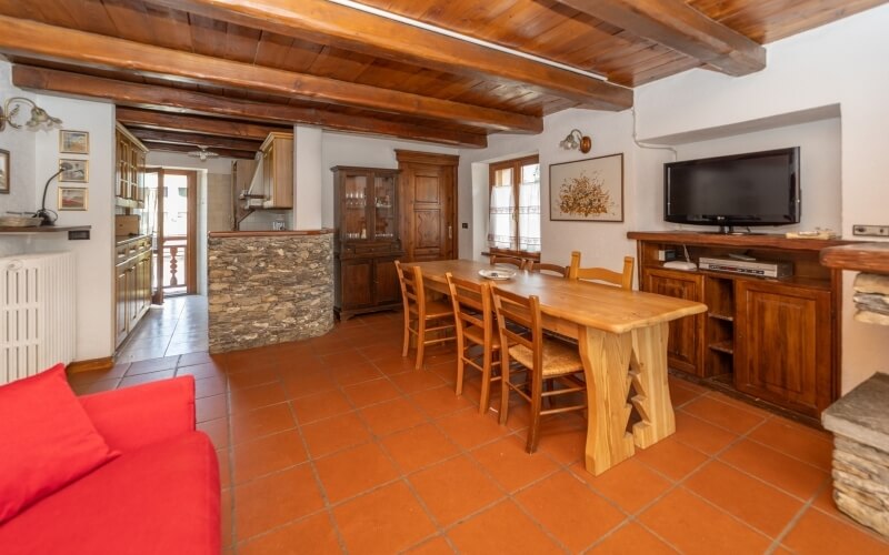 Rustic room with dining area, kitchen, and living space; features red couch, wooden table, and terracotta tile flooring.