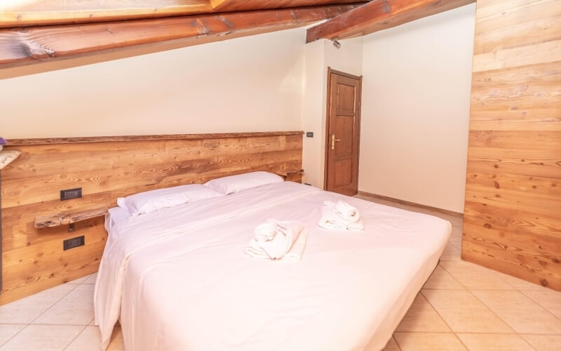 Camera serena con parete in legno, letto con coperta bianca e asciugamani, pavimento in piastrelle beige e travi a vista.