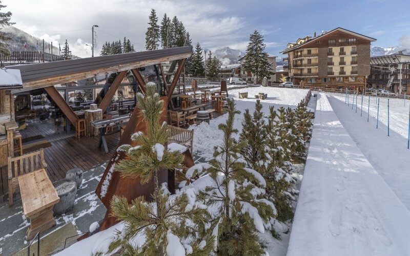 Area esterna di ristorante con tavoli, panchine e alberi di pino innevati.