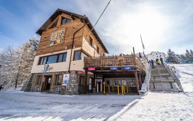 Exterior of a ski resort building with wooden and brick design, colorful banners, deck, and snowy surroundings.