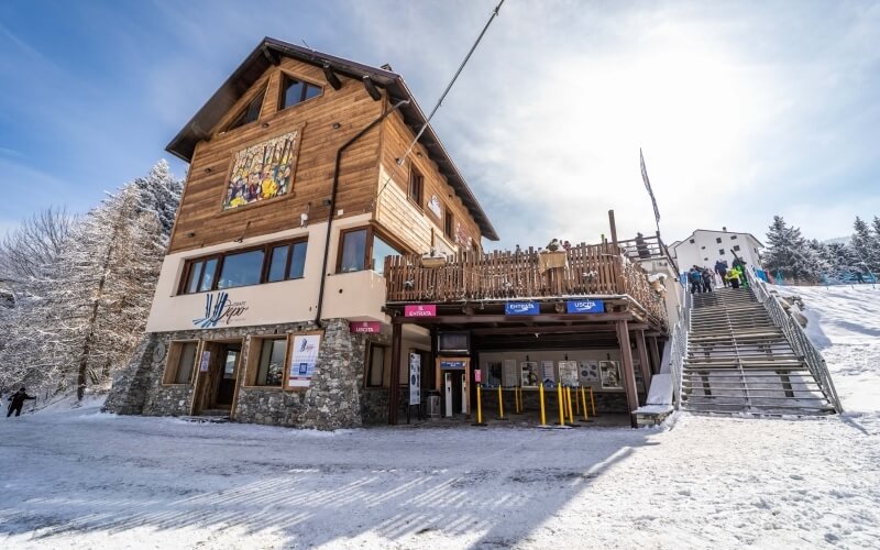 Two-story building on a snowy hill with stone and wood accents, surrounded by trees under a blue, cloudy sky.