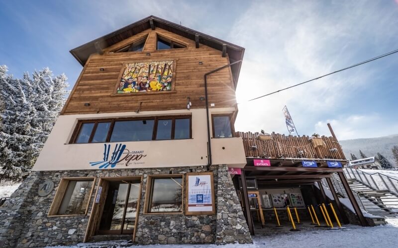 Building with stone foundation and wooden upper section, "SMART YOGA" sign, snowy landscape, and cable car poles in foreground.