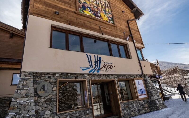 Two-story building with "SMART Depo" sign, stone base, wood accents, snowy mountains in background, and people nearby.