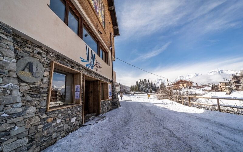 Scena invernale serena con edificio in pietra e legno, logo "Alpes", montagne innevate e cielo blu.