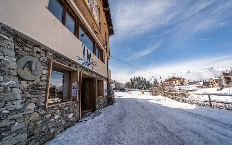 Exterior of a rustic building with a stone wall, wooden window, "Alpes" sign, snowy road, and clear blue sky.