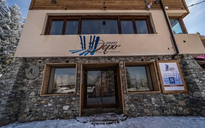 Two-story building with stone base, "SMART Depo" sign, snowy ground, and trees against a blue sky.