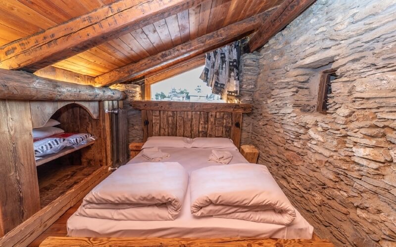Cozy cabin room with a bed, loft area, wooden beams, stone wall, and natural light from a large window.