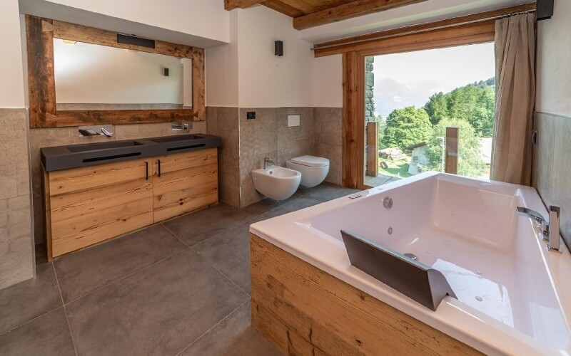 Bagno moderno con vasca bianca, due WC, lavabo e grande finestra con vista su alberi e montagne.