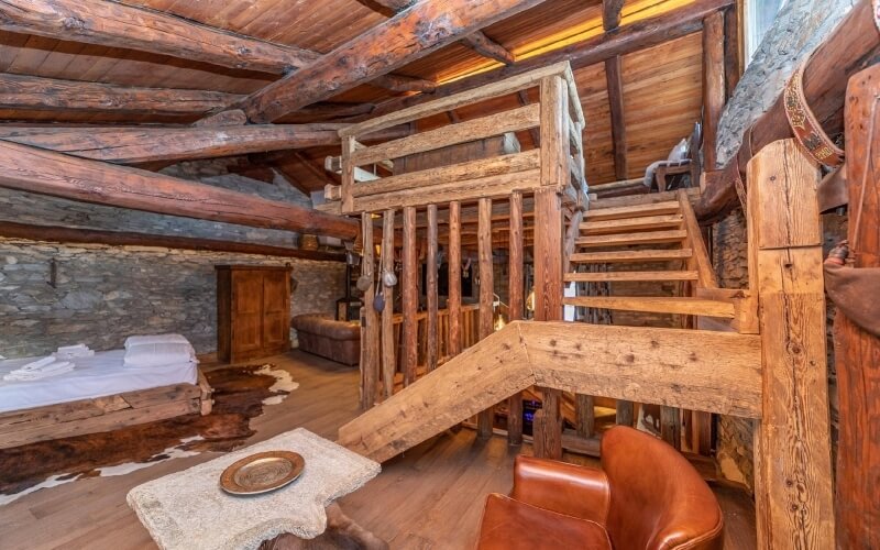 Rustic two-story room with wooden beams, stone walls, staircase, leather chairs, cowhide rug, and cozy furniture.