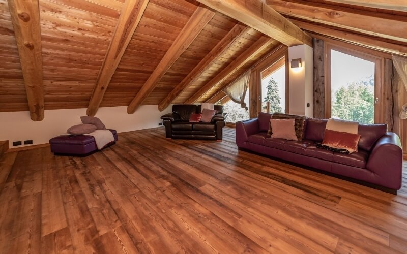 Una stanza spaziosa con soffitto inclinato, finestre grandi, due divani in pelle marrone e un pouf viola con cuscini colorati.