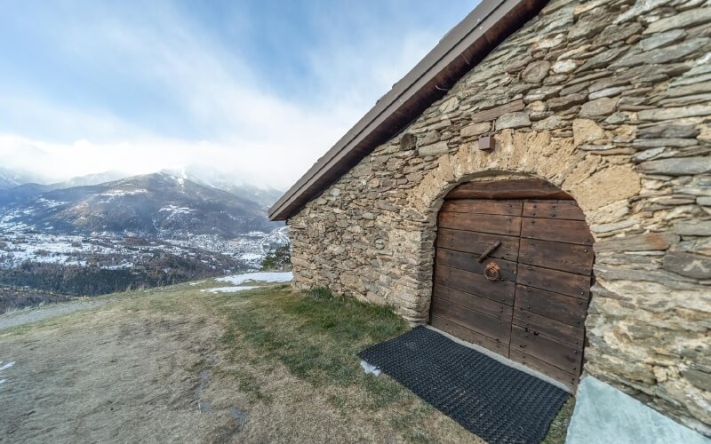 Stone building with wooden door in a snowy mountainous landscape under a blue sky, with a distant village visible.