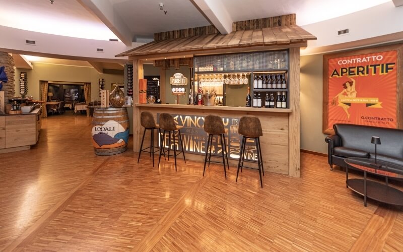 Modern bar area with light wood bar, leather stools, gray sofa, orange poster, and a doorway to a dining area.