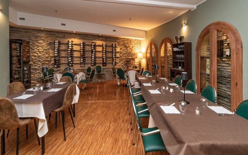 Formal dining room with long tables, green-backed chairs, a stone wall, wine racks, and a bar area, creating an elegant atmosphere.