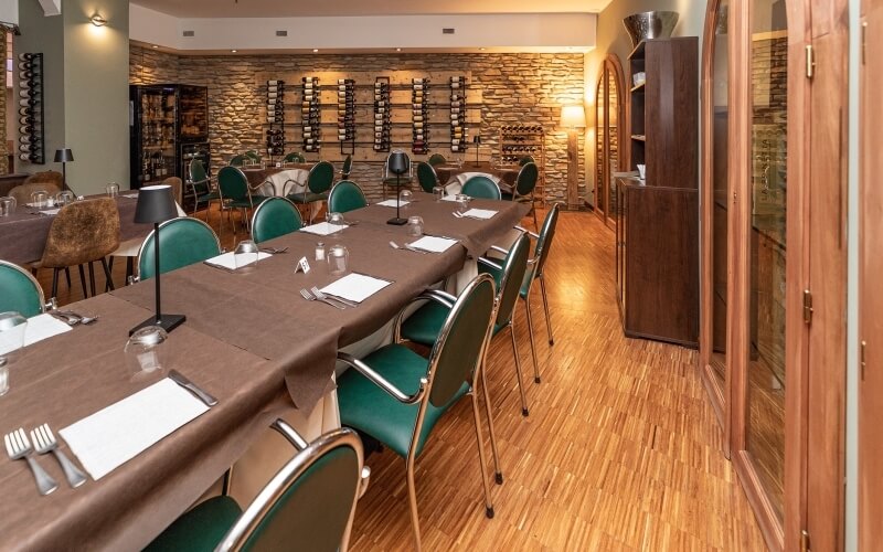 A restaurant dining room with long tables, brown tablecloths, green chairs, and a stone wall with wine racks, creating an elegant atmosphere.