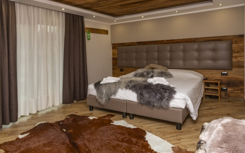 Hotel room featuring a large bed with gray fur pillows, wooden nightstand, and neutral decor in beige and brown tones.