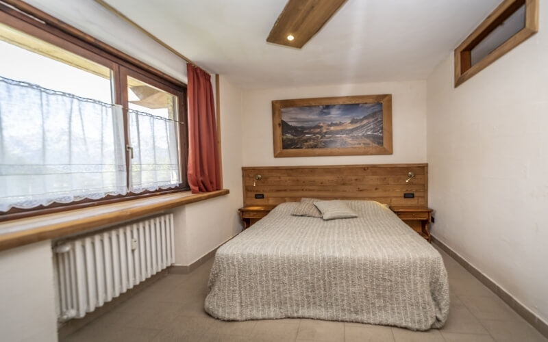 Camera serena e accogliente con letto matrimoniale, pareti bianche, accenti in legno e luce naturale da una finestra.