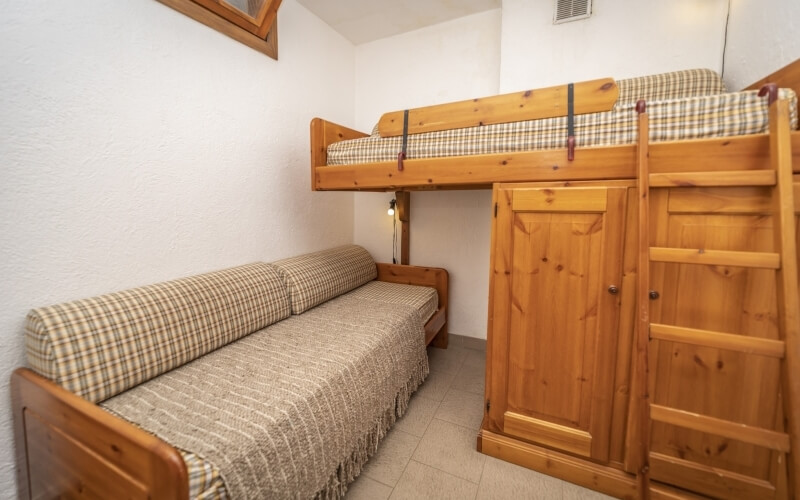 Una stanza piccola con un letto a castello in legno chiaro, un divano e pareti bianche, che trasmette un'atmosfera accogliente.