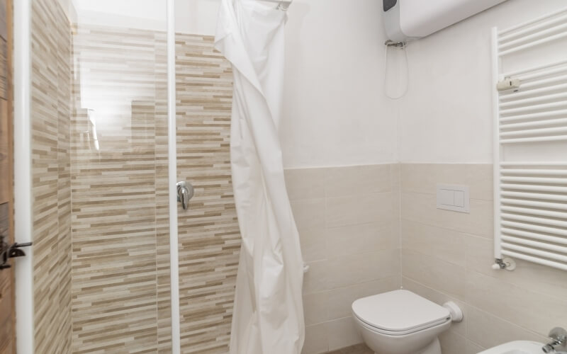 Bagno pulito con doccia in vetro, WC bianco, radiatore e porta in legno con maniglia argentata.