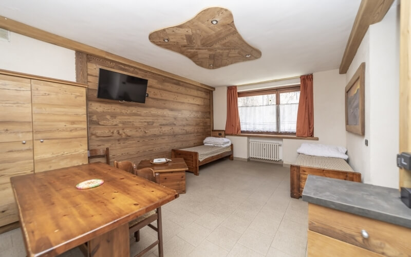 Stanza rustica con pareti in legno, letti a due piazze, tavolo in legno, TV a muro e finestre con tende rosse.