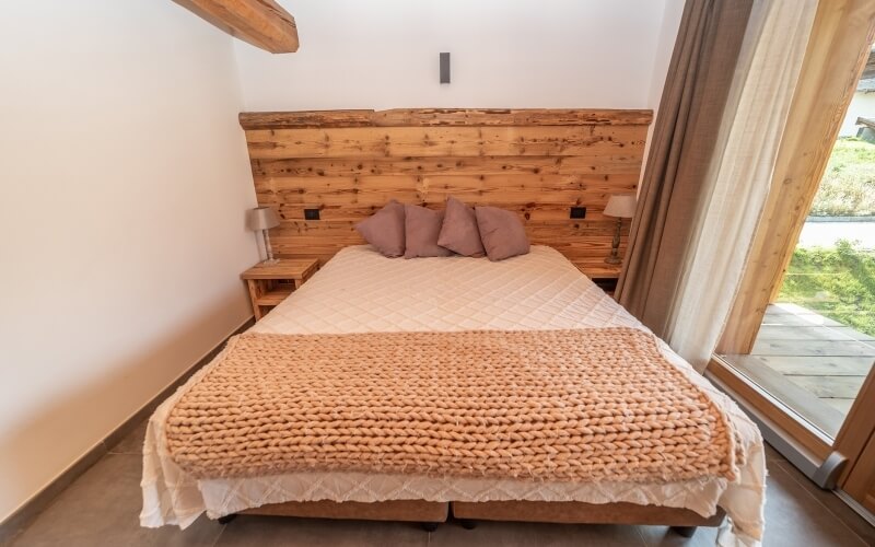 Camera da letto serena con letto, testiera in legno, coperta bianca, cuscini rosa e luce naturale da una grande finestra.
