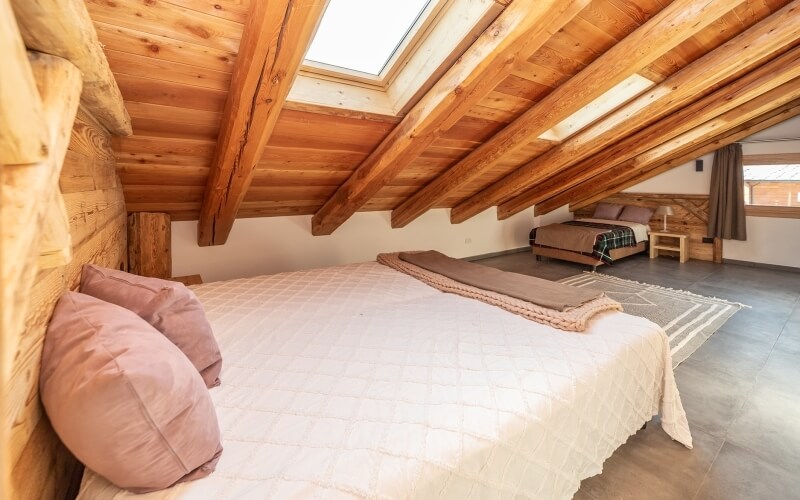Camera spaziosa con travi in legno, letti bianchi e marroni, luce naturale da un lucernario e finestre, atmosfera accogliente.