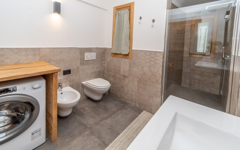 Bagno pulito con area lavanderia, tavolo in legno, lavatrice, bidet, toilet, specchio e doccia con porta in vetro.