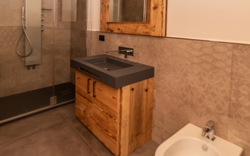 Bagno moderno con doccia, lavabo, specchio e bidet, caratterizzato da piastrelle chiare e pavimento scuro.