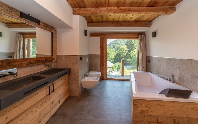 Bagno moderno con finestra panoramica, specchio grande, due lavandini, vasca, e vista su un paesaggio sereno.