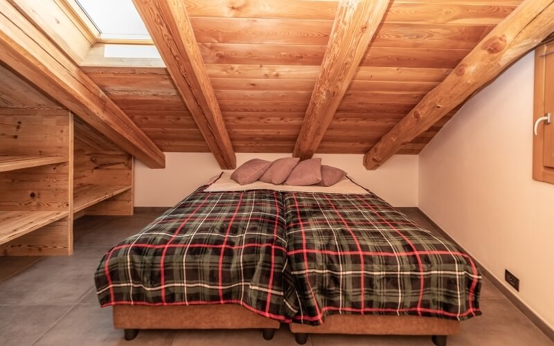 Camera accogliente con letto a parete, coperta a quadri, travi a vista, finestra e pavimento in piastrelle grigie.