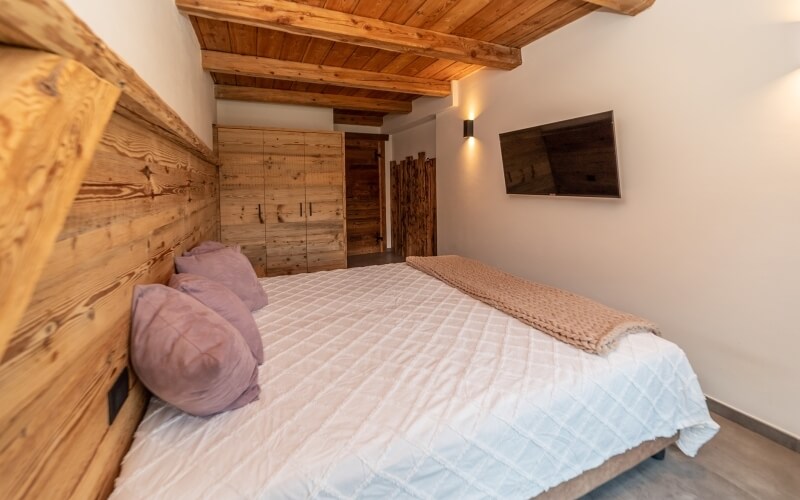 Camera da letto serena con letto bianco, testiera in legno, TV a parete e toni caldi di legno e rosa.