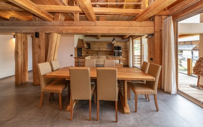 Una spaziosa sala da pranzo con un grande tavolo in legno chiaro, sedie beige e una cucina rustica visibile sullo sfondo.