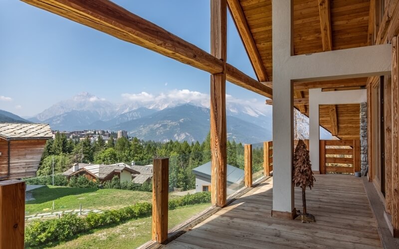 Wooden house with a deck overlooking trees and mountains under a clear blue sky, creating a serene and tranquil scene.