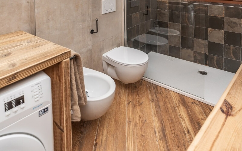 Compact bathroom with a toilet, shower, washing machine, and wooden counters; features glass tile and light wood flooring.