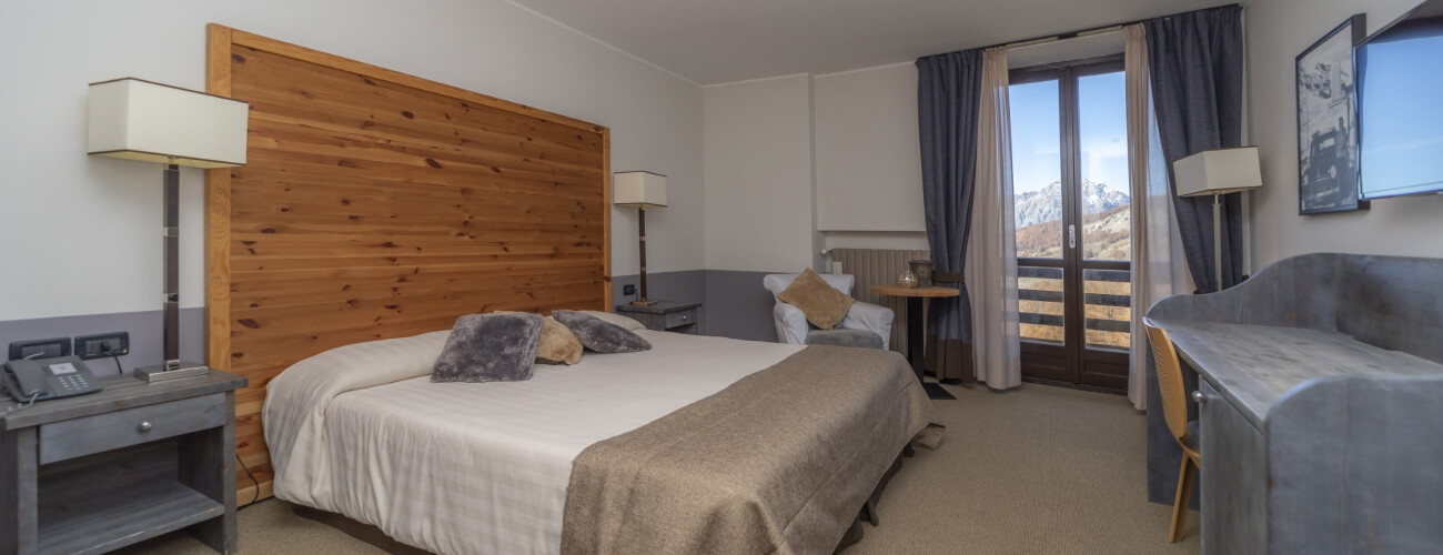 Camera d'hotel accogliente con letto grande, scrivania grigia, divano e vista sulle montagne attraverso una finestra a golfo.