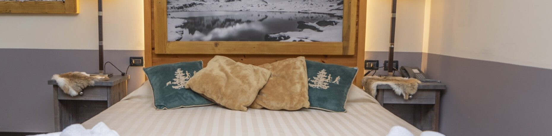 Cozy bedroom with a bed, decorative pillows, nightstands with lamps, and a snowy landscape picture above.