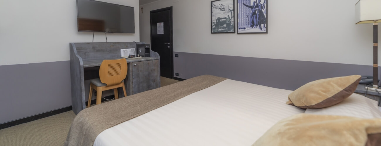 Minimalist hotel room featuring a bed, desk, television, and decorative pictures, with a neutral color scheme.